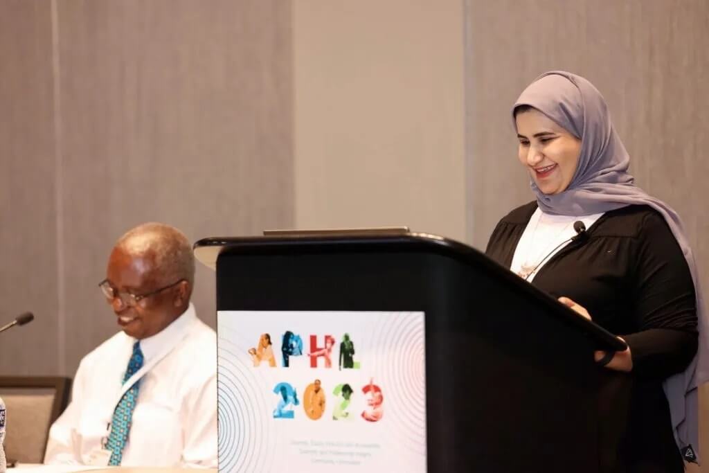A speaker smiles while standing at a podium and another speaker sits alongside.