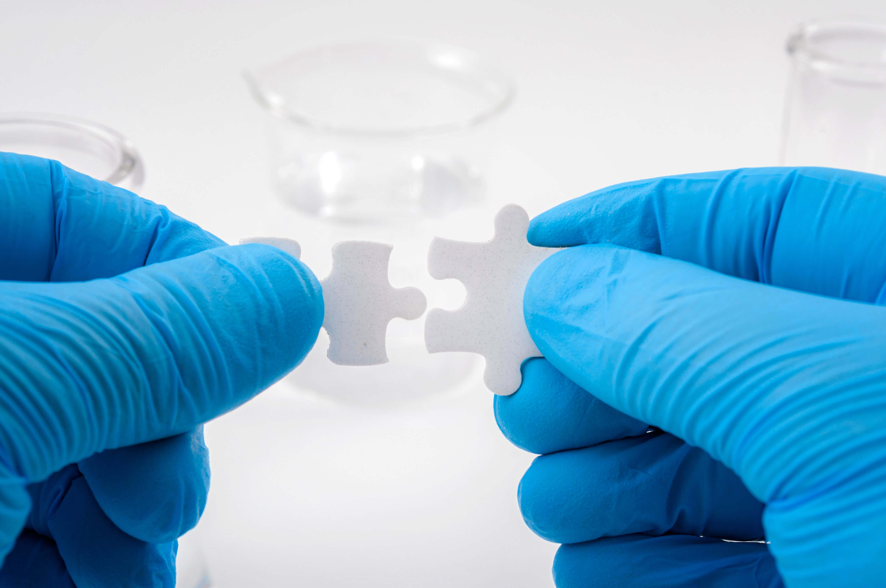 Two hands wearing blue gloves connecting two puzzle pieces, beaker with clear liquid in background